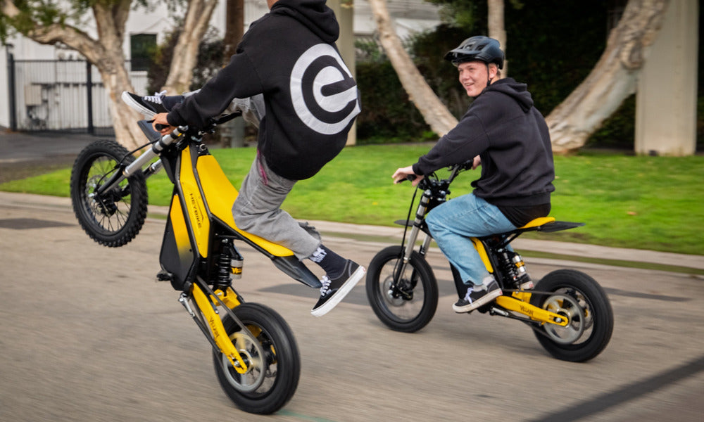 Two riders on yellow Heybike Sports V1 electric dirt bikes, one doing a wheelie on a neighborhood street while the other rides beside him.