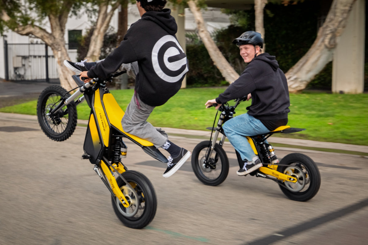 Two riders on yellow Heybike Sports V1 electric dirt bikes, one doing a wheelie on a neighborhood street while the other rides beside him.
