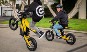 Two riders on yellow Heybike Sports V1 electric dirt bikes, one doing a wheelie on a neighborhood street while the other rides beside him.